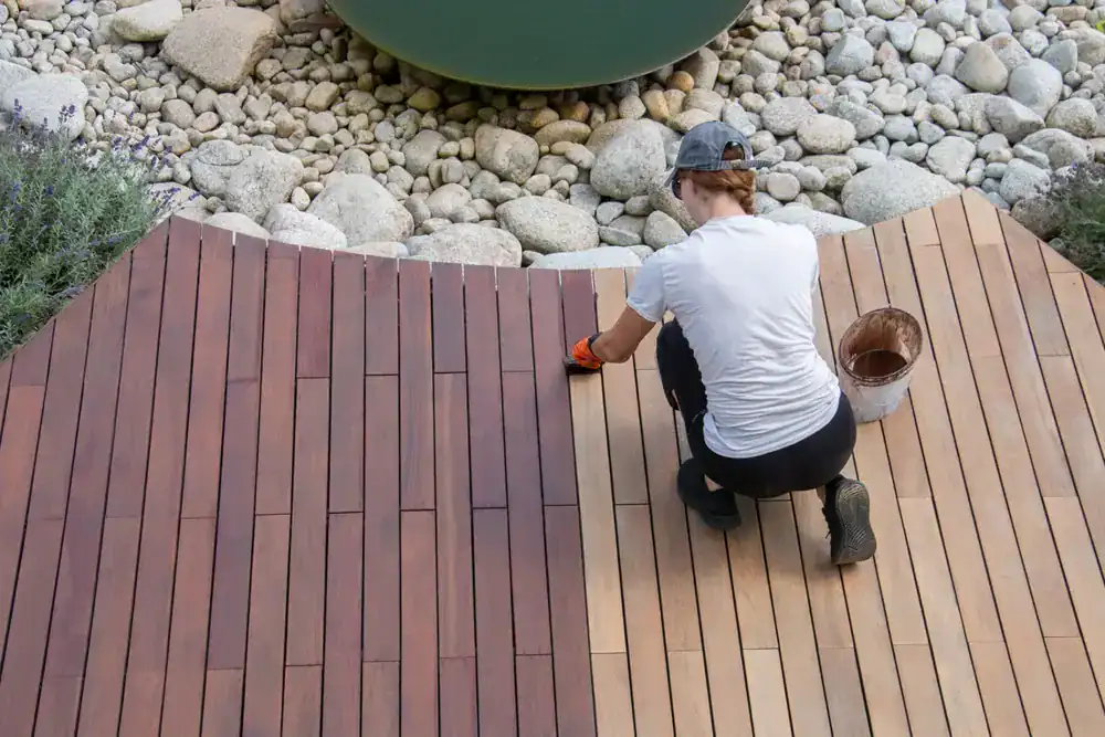 A person kneels on a wooden deck in MI, staining the light-colored planks to a darker shade, with Painters Macomb & Oakland County’s supplies and a bucket of stain nearby, and round stones visible in the background.