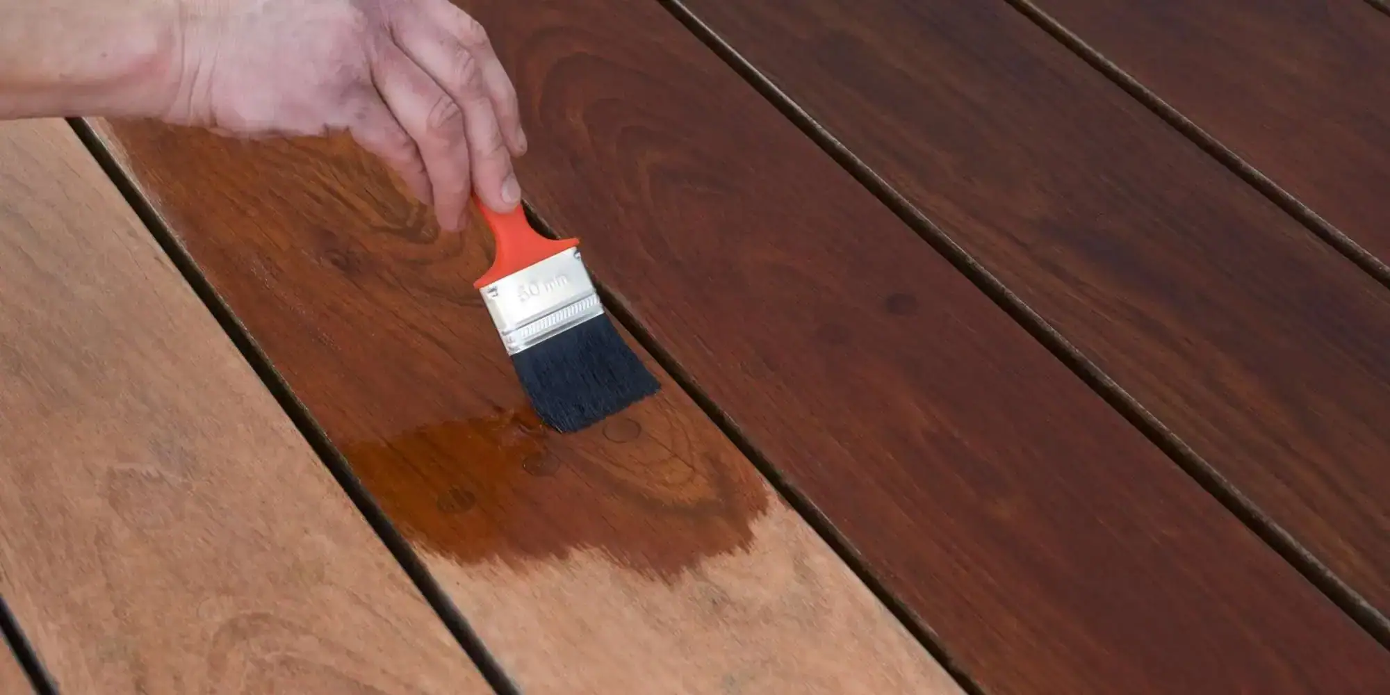 A hand uses a paintbrush to apply a dark wood stain to a wooden deck, revealing a clear contrast between treated and untreated sections&mdash;expertly done by MI, Painters Macomb & Oakland County.