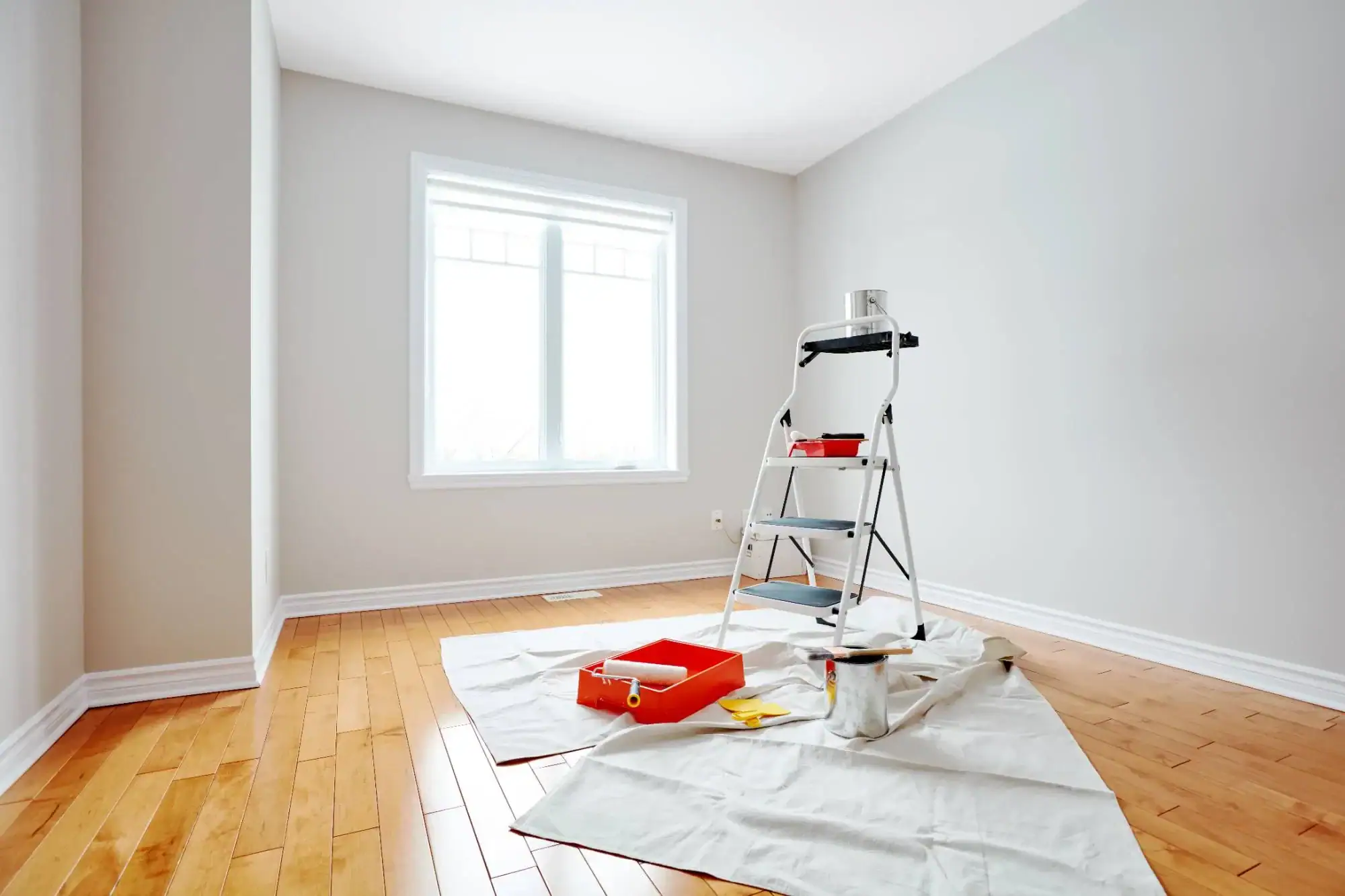 A bright, empty room with light hardwood floors and pale walls. A window lets in natural light. A ladder, paint cans, and supplies from Painters Macomb & Oakland County, MI, rest on a drop cloth, suggesting the room is being painted.