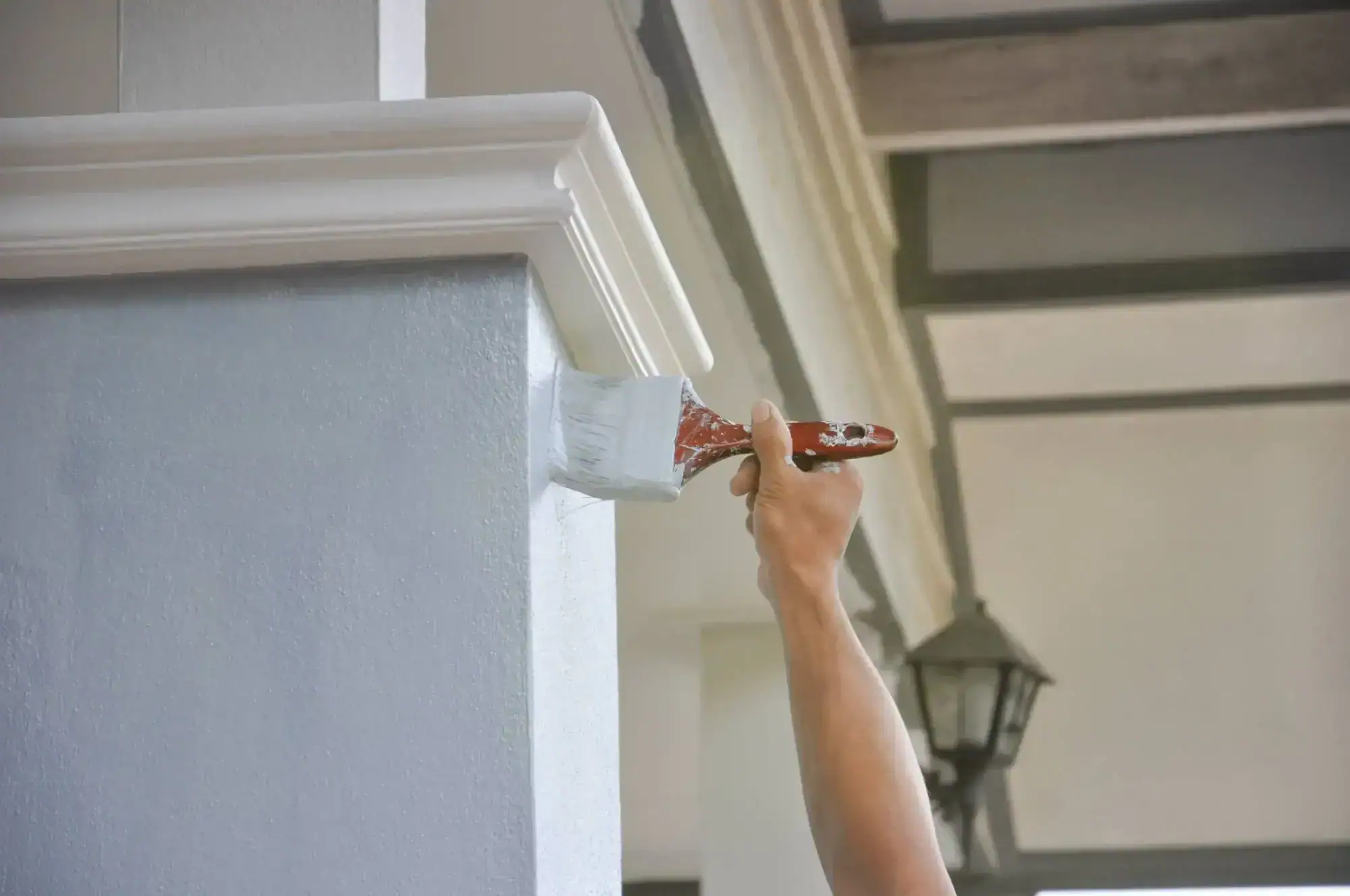 A person&rsquo;s hand paints the edge of a light gray exterior column with white paint using a paintbrush. In the background, part of a building and a hanging outdoor lantern are seen&mdash;showcasing skilled work by painters in Macomb & Oakland County, MI.
