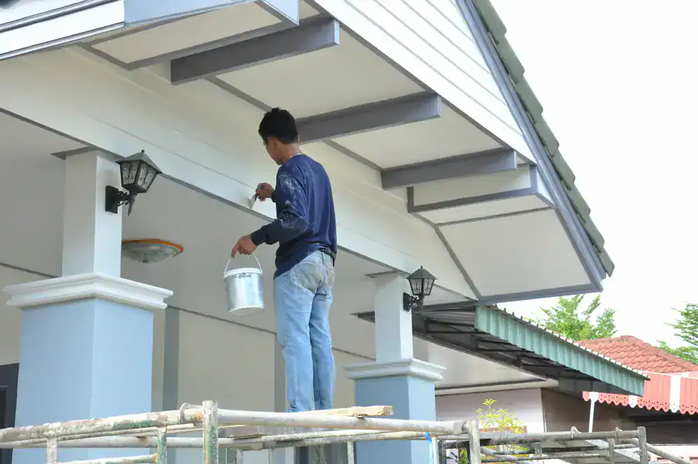 A person in jeans and a long-sleeve shirt stands on scaffolding, painting the exterior trim of a house. Painters Macomb & Oakland County, MI specialize in homes with white and light blue pillars and sloped roofs like this one.