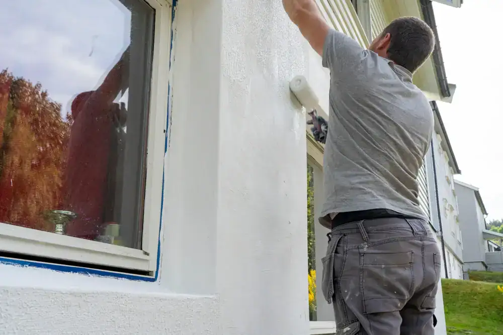 A person in a gray shirt and work pants is painting the exterior wall of a house white, standing next to a window with blue painter’s tape—just like the pros at Painters Macomb & Oakland County, MI.
