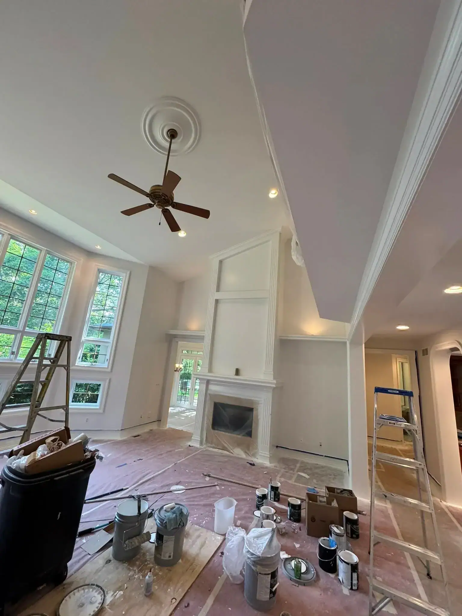 A bright living room with high ceilings, white walls, and large windows shows painters from Macomb & Oakland County, MI at work. Ladders, paint cans, and supplies are scattered as renovations are underway, with the floor covered for protection.
