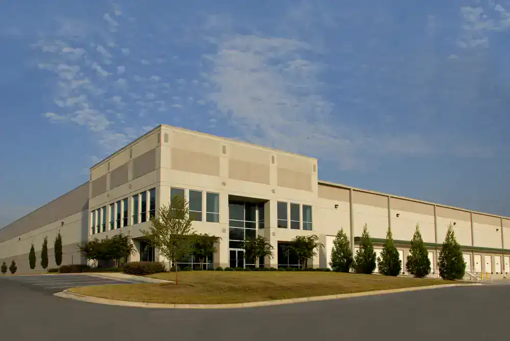 A large, modern warehouse building with tall windows and beige walls, freshly painted by expert painters in Macomb & Oakland County, MI, sits amid neatly trimmed trees and an empty parking lot under a partly cloudy sky.