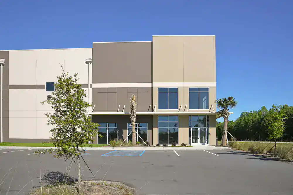 A modern, beige industrial building with large windows is shown under a clear blue sky. This property, ideal for painters in Macomb & Oakland County, MI, features small trees and empty parking spaces—including a marked accessible spot—out front.