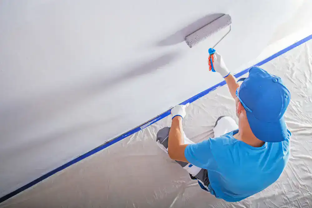 A person from Painters Macomb & Oakland County, MI, wearing a blue shirt, cap, and white gloves, uses a paint roller to paint a wall white. Protective sheets and tape cover the floor and edges to prevent spills.