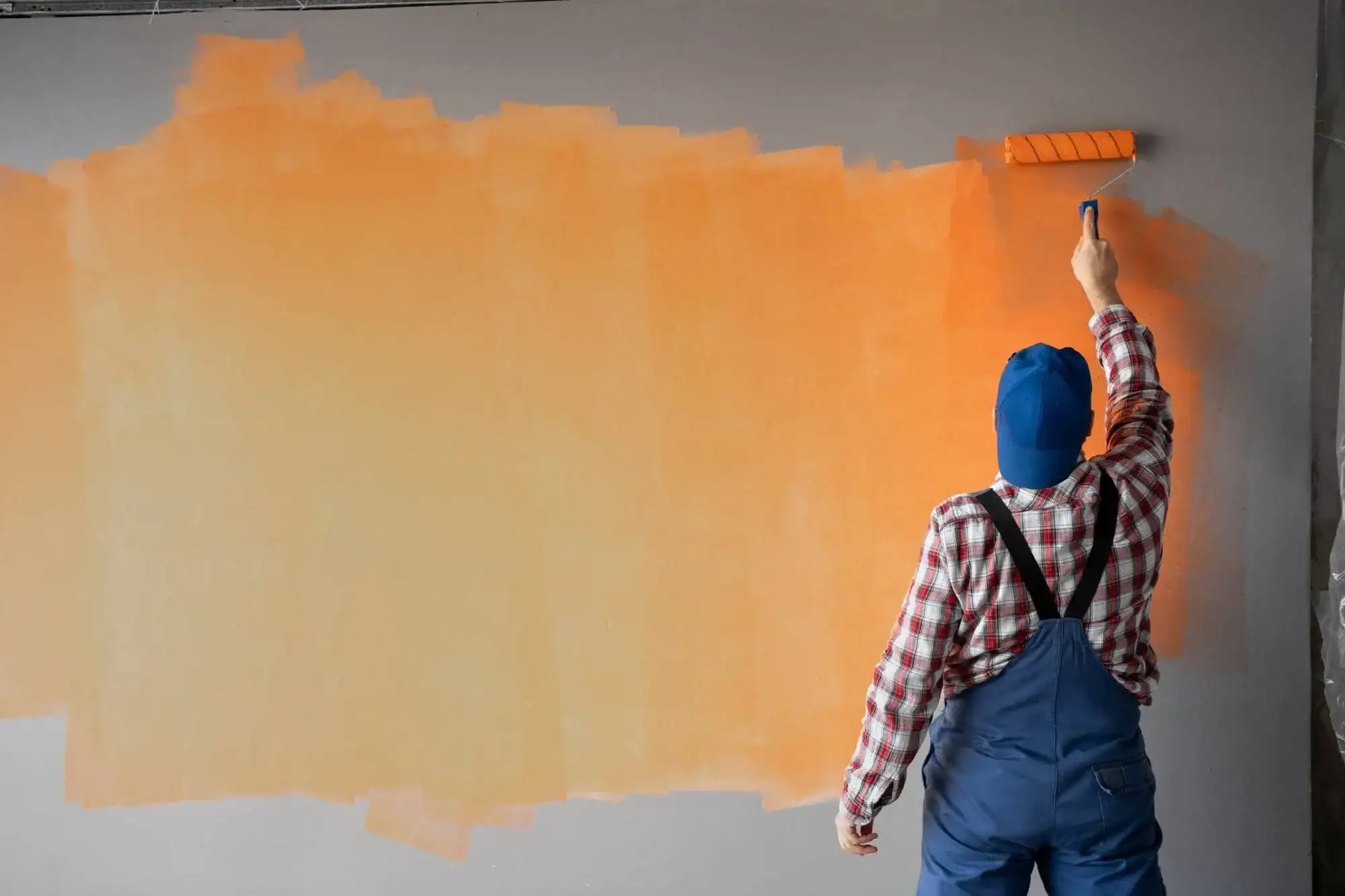 A person wearing a blue beanie, plaid shirt, and blue overalls is painting a gray wall orange with a paint roller. MI Painters Macomb & Oakland County professionals often achieve such impressive coverage on large walls.