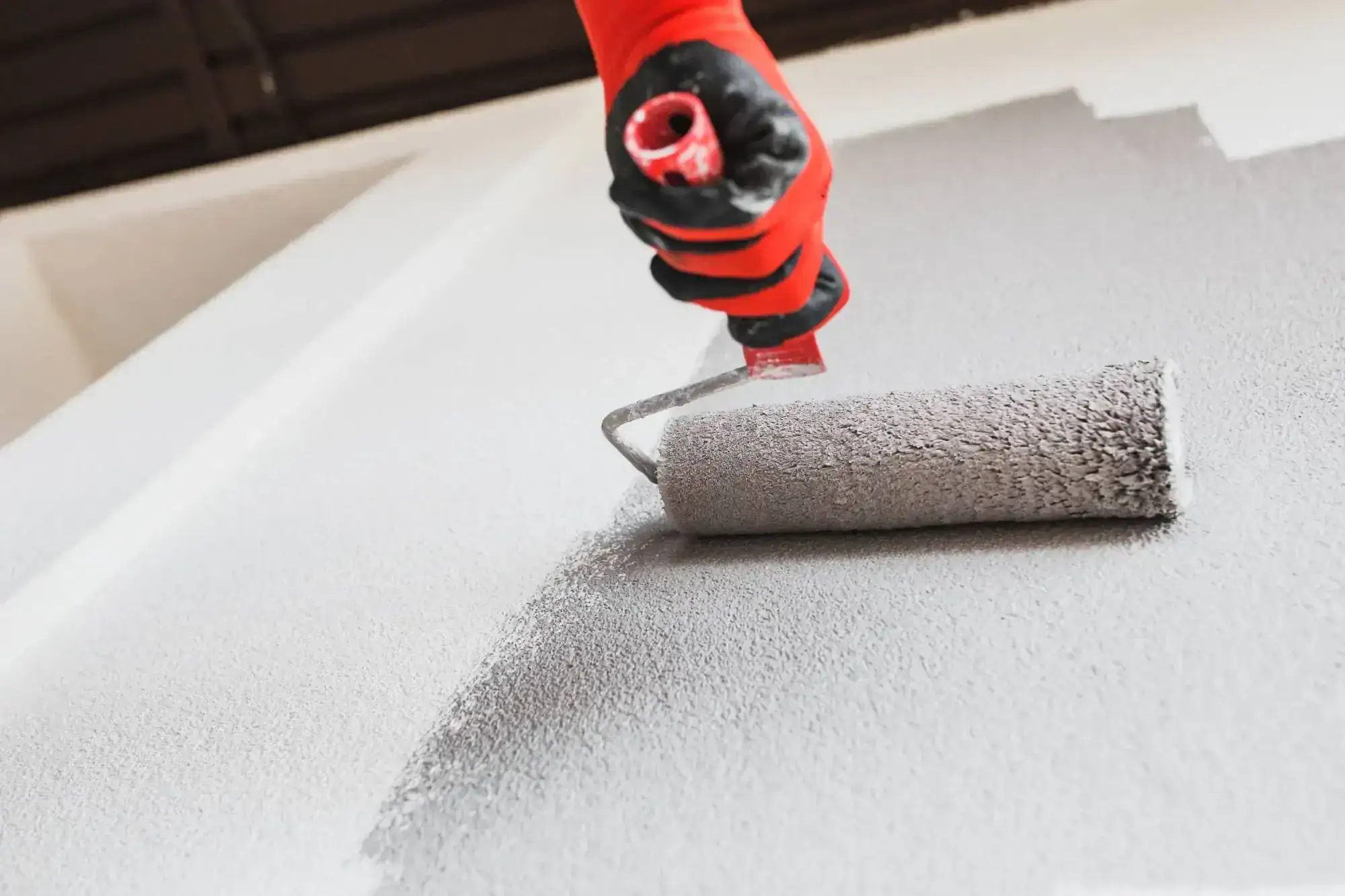 A person wearing a red glove paints a wall with a grey roller, applying fresh light grey paint. The roller moves upward, leaving a smooth finish on the textured surface&mdash;quality work from Painters Macomb & Oakland County, MI.