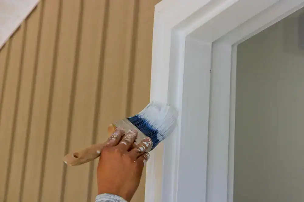 A hand holding a paintbrush is painting the trim of a window white. The background shows tan vertical siding, showcasing the skilled work of Painters Macomb & Oakland County, MI.