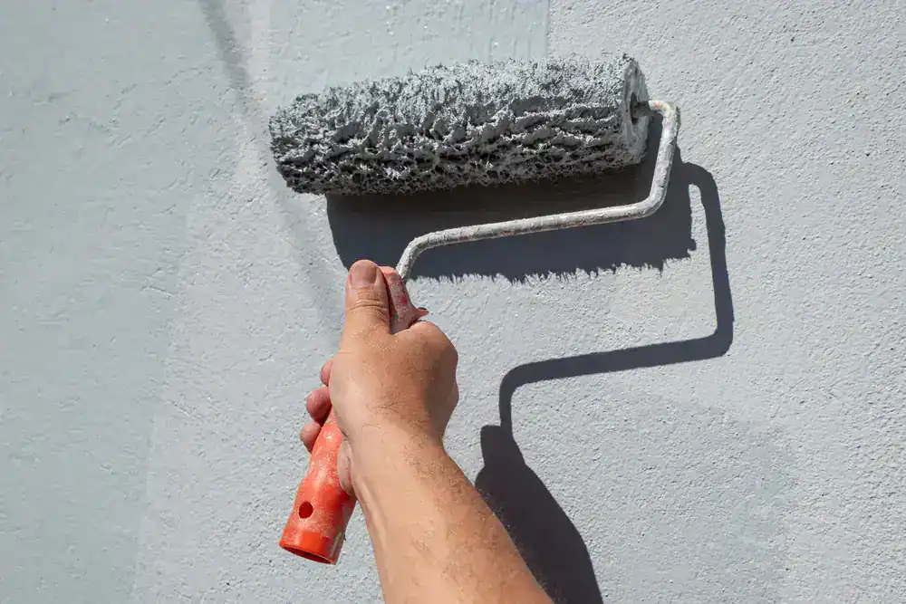A hand holds an orange-handled paint roller, applying light gray paint to a smooth wall in MI. The roller is covered with paint, and the shadow of the roller is visible on the wall&mdash;expert work by Painters Macomb & Oakland County.
