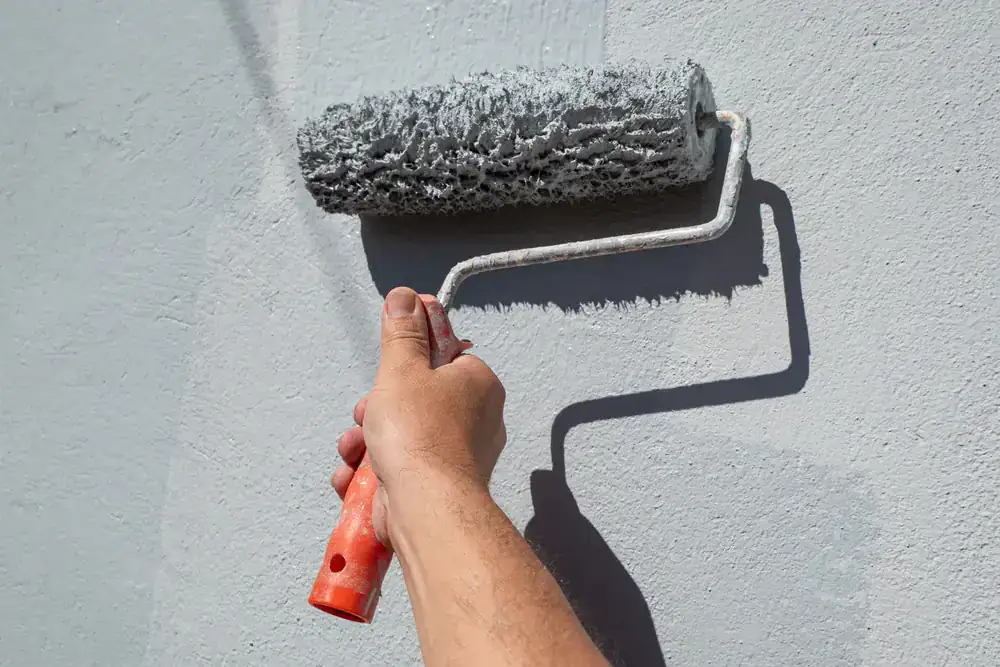 A hand holding an orange-handled paint roller applies light gray paint to a wall in MI, creating a smooth, even coat. The roller casts a shadow on the painted surface&mdash;a signature touch of Painters Macomb & Oakland County.