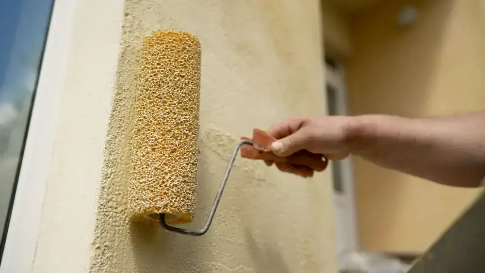 A hand uses a yellow textured paint roller to apply beige paint to an exterior wall near a window, showcasing the careful work of MI Painters Macomb & Oakland County.