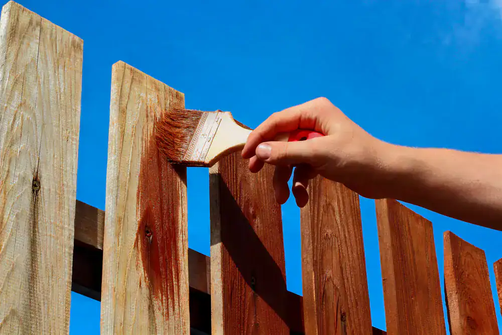 A hand holding a paintbrush applies brown stain to a wooden fence under a clear blue sky, with some boards freshly coated and others still unpainted—a classic scene for MI Painters Macomb & Oakland County.