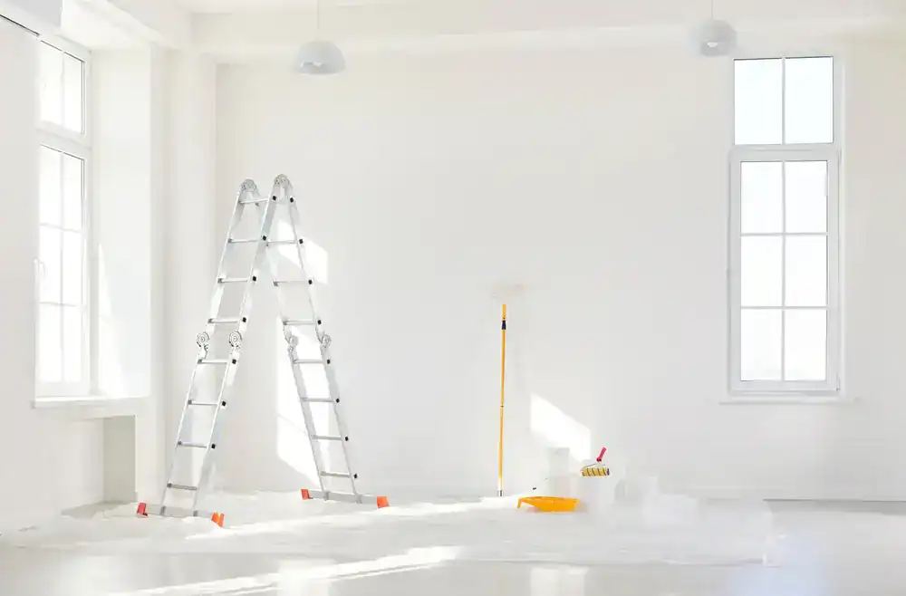 A bright, empty room with white walls and tall windows features a folding ladder, a paint roller on an extension pole, and painting supplies on a plastic-covered floor—ready for MI Painters Macomb & Oakland County to transform the space.