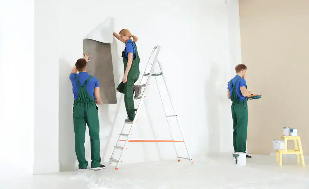 Three people wearing green overalls are redecorating a room; Painters Macomb & Oakland County, MI, with one on a ladder peeling wallpaper, another painting a wall, and the third preparing supplies on a small stool.