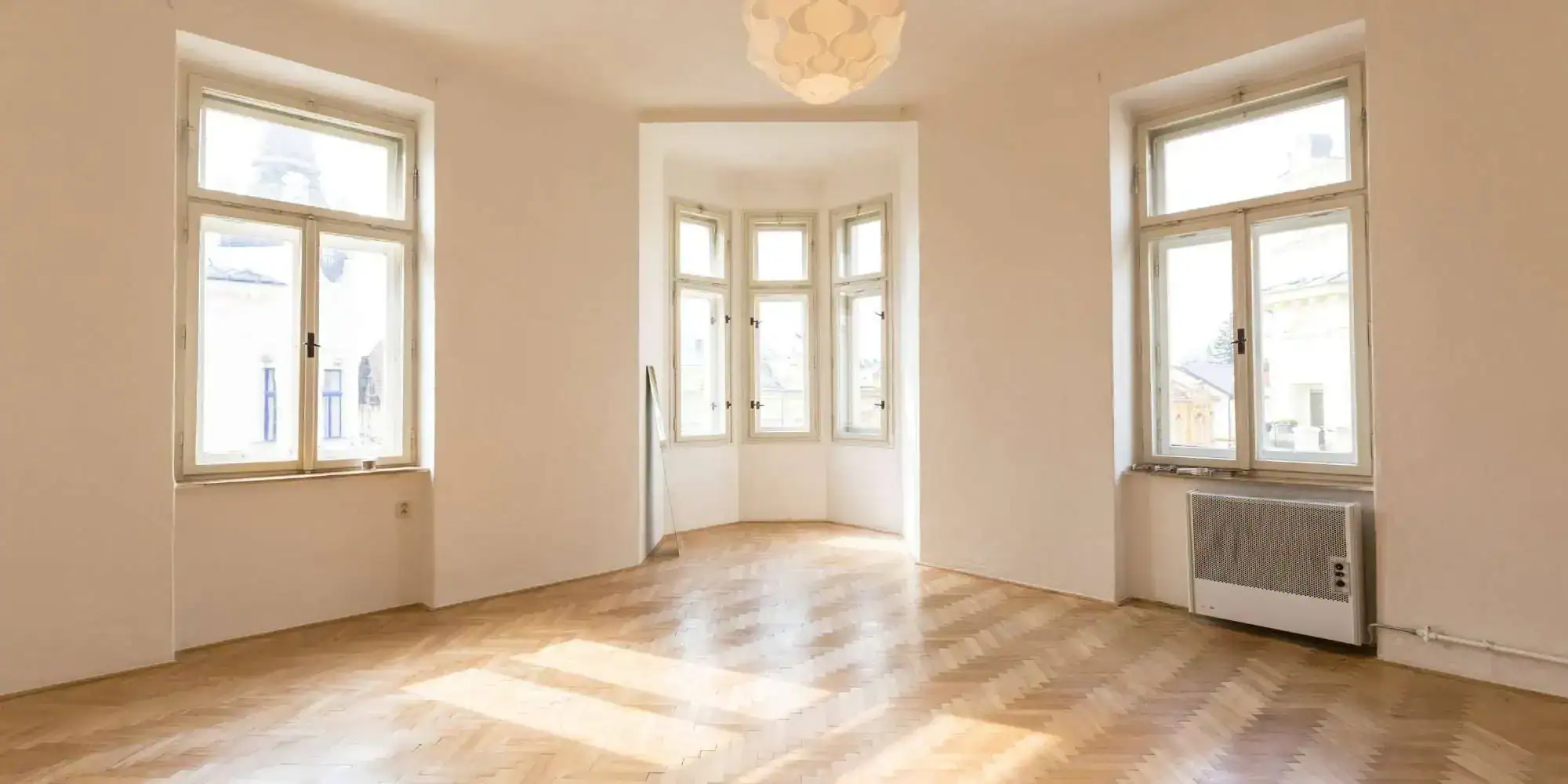 Bright, empty room with large windows, light-colored walls, wooden parquet flooring, a decorative ceiling light, and a bay window area letting in natural light—a perfect canvas for MI Painters Macomb & Oakland County to transform.