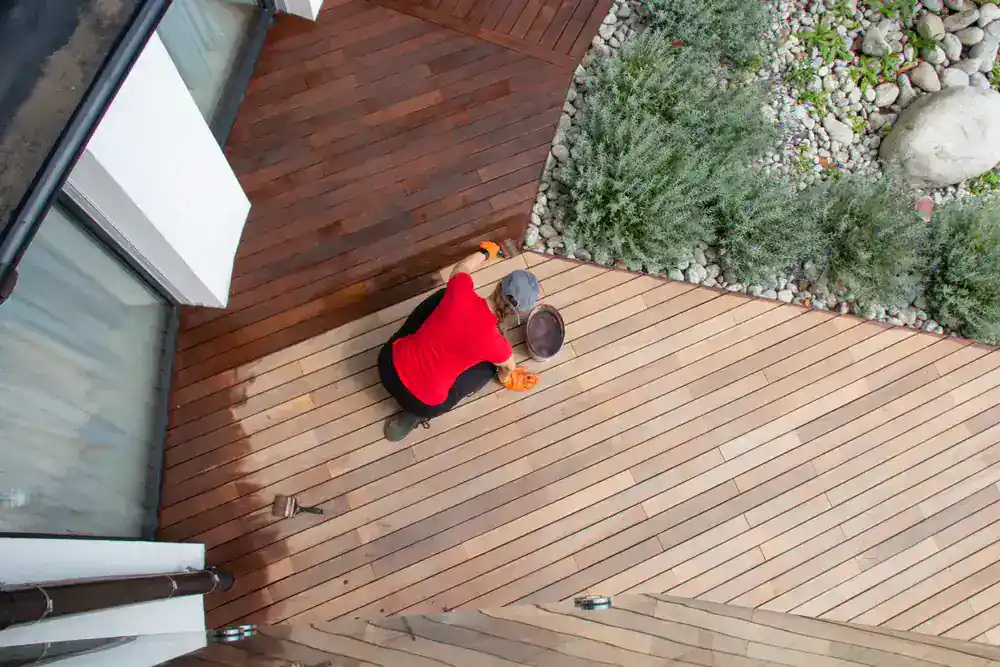 A person wearing a red shirt and orange gloves is staining or cleaning a wooden deck in MI, kneeling with a bucket and cloth near green plants and rocks&mdash;a scene captured from above, perfect for Painters Macomb & Oakland County inspiration.