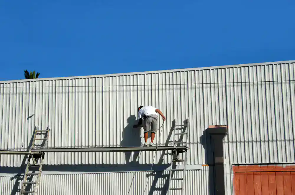 A person in a white shirt and shorts stands on scaffolding, painting or working on the side of a large, white corrugated metal building under a clear blue MI sky&mdash;just another day for skilled Painters Macomb & Oakland County.