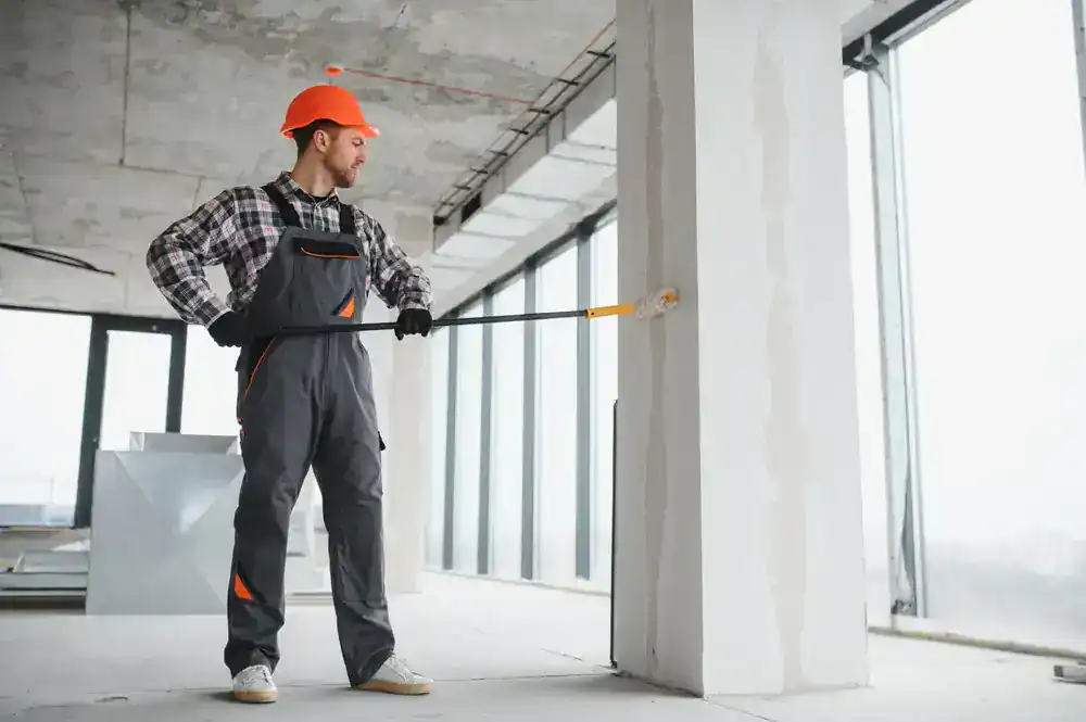 A construction worker wearing a hard hat and overalls uses a paint roller to paint a large white pillar in a modern, unfinished building with large windows in MI, showcasing the skill of Painters Macomb & Oakland County.