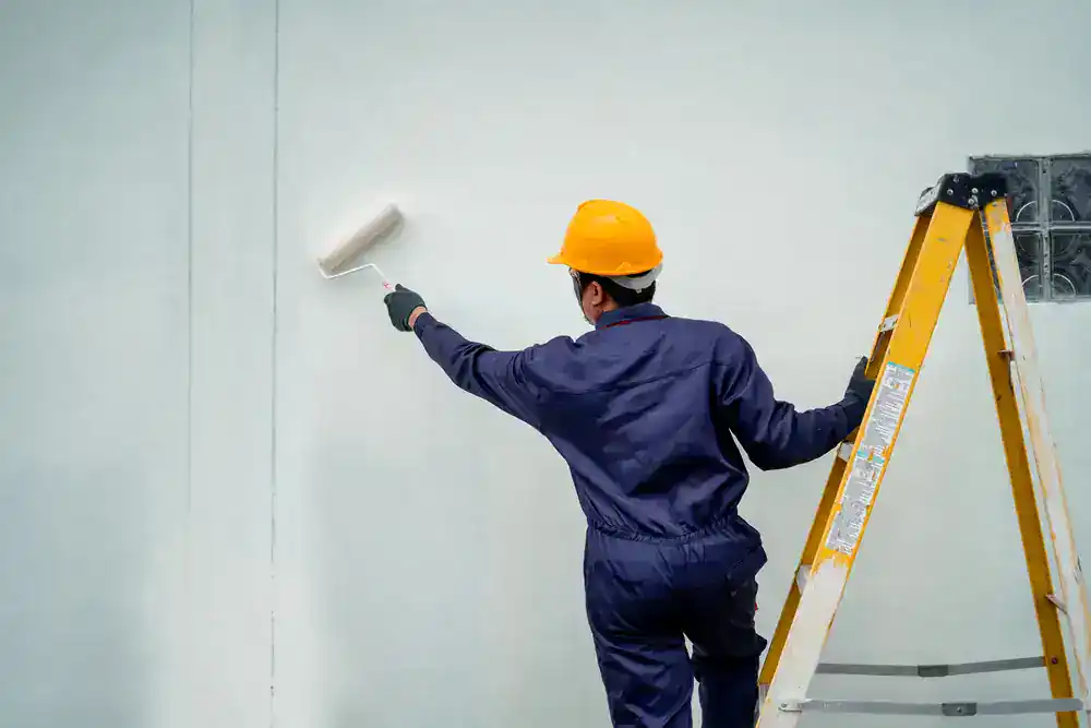 A person wearing a yellow hard hat and navy coveralls uses a paint roller to paint a white wall, standing next to a yellow stepladder&mdash;representing Painters Macomb & Oakland County, MI.
