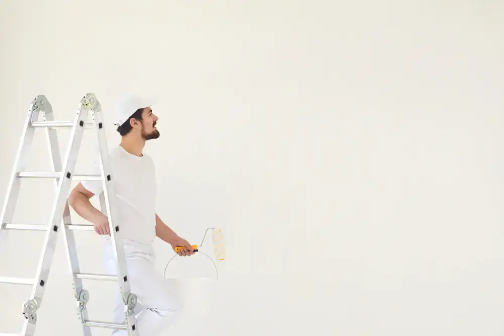 A man dressed in white work clothes and cap stands beside a ladder, smiling and holding a paint scraper, representing Painters Macomb & Oakland County, MI, with a blank white wall in the background.
