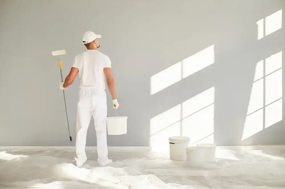 A person dressed in white holds a paint roller and bucket, standing before a light gray wall with sunlight pouring through large windows. Three more paint buckets rest on a drop cloth—ready for Painters Macomb & Oakland County, MI to get started.
