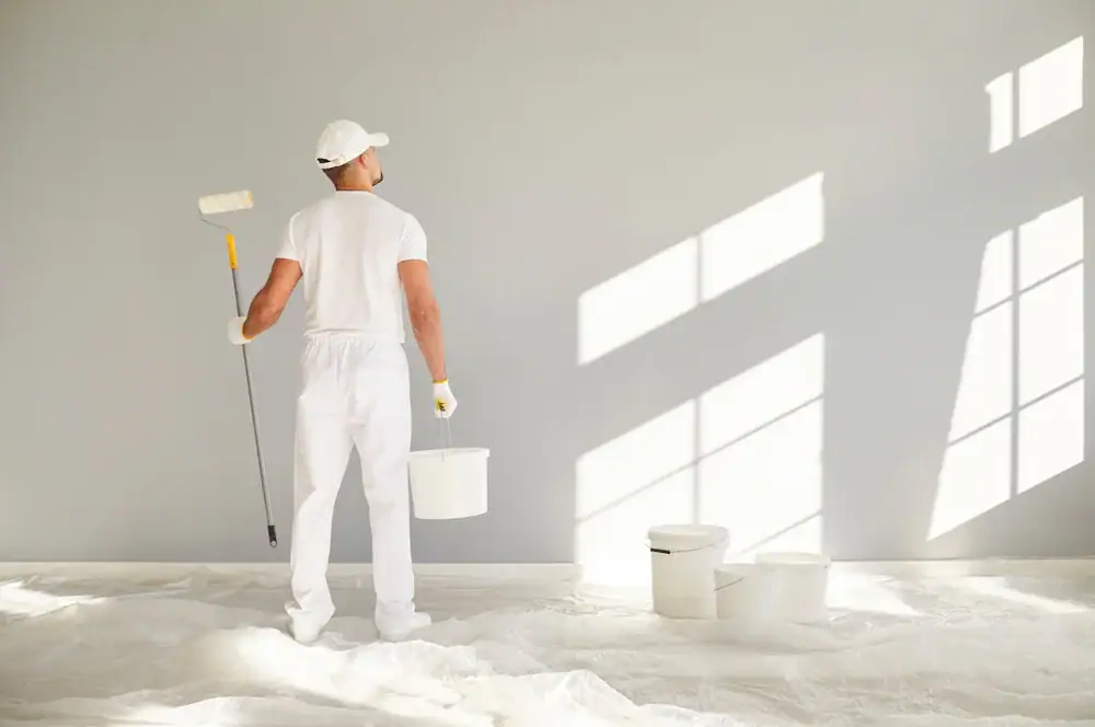 A person dressed in white work clothes and cap, like painters in Macomb & Oakland County, MI, holds a paint roller and bucket, standing before a light gray wall as sunlight streams through windows; paint buckets rest on a tarp-covered floor.