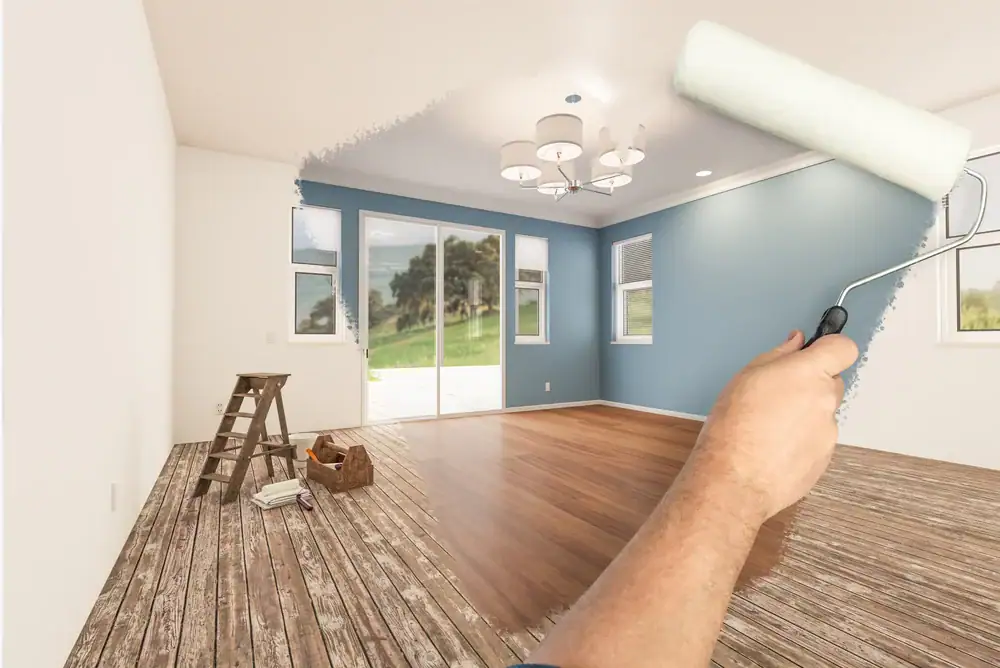 A hand uses a paint roller to transform a plain white room into a modern living space with blue walls, wood flooring, and new lighting. MI Painters Macomb & Oakland County supplies and a ladder sit nearby, ready for the renovation.