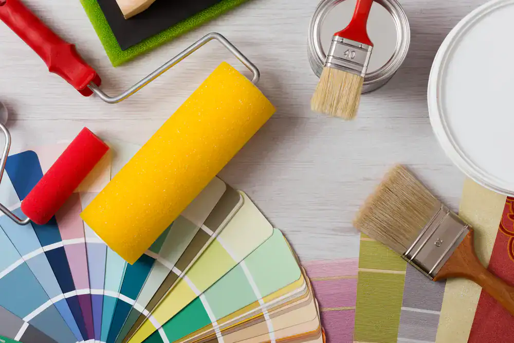 A yellow paint roller, paintbrushes, paint cans, and a color swatch palette are arranged on a light wooden surface, showcasing tools and choices for interior painting by Painters Macomb & Oakland County, MI.