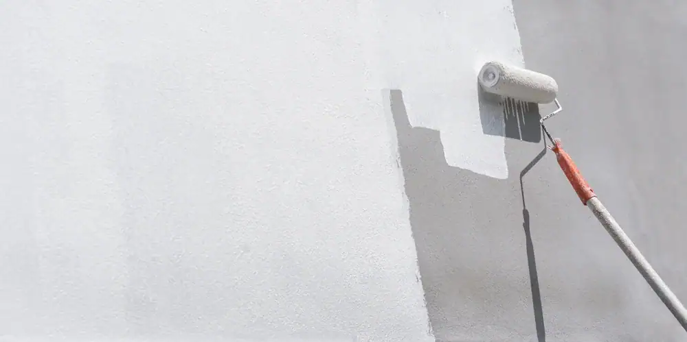 A paint roller with a long handle is being used to paint a white wall, covering a darker section with fresh white paint—just another day for Painters Macomb & Oakland County, MI.