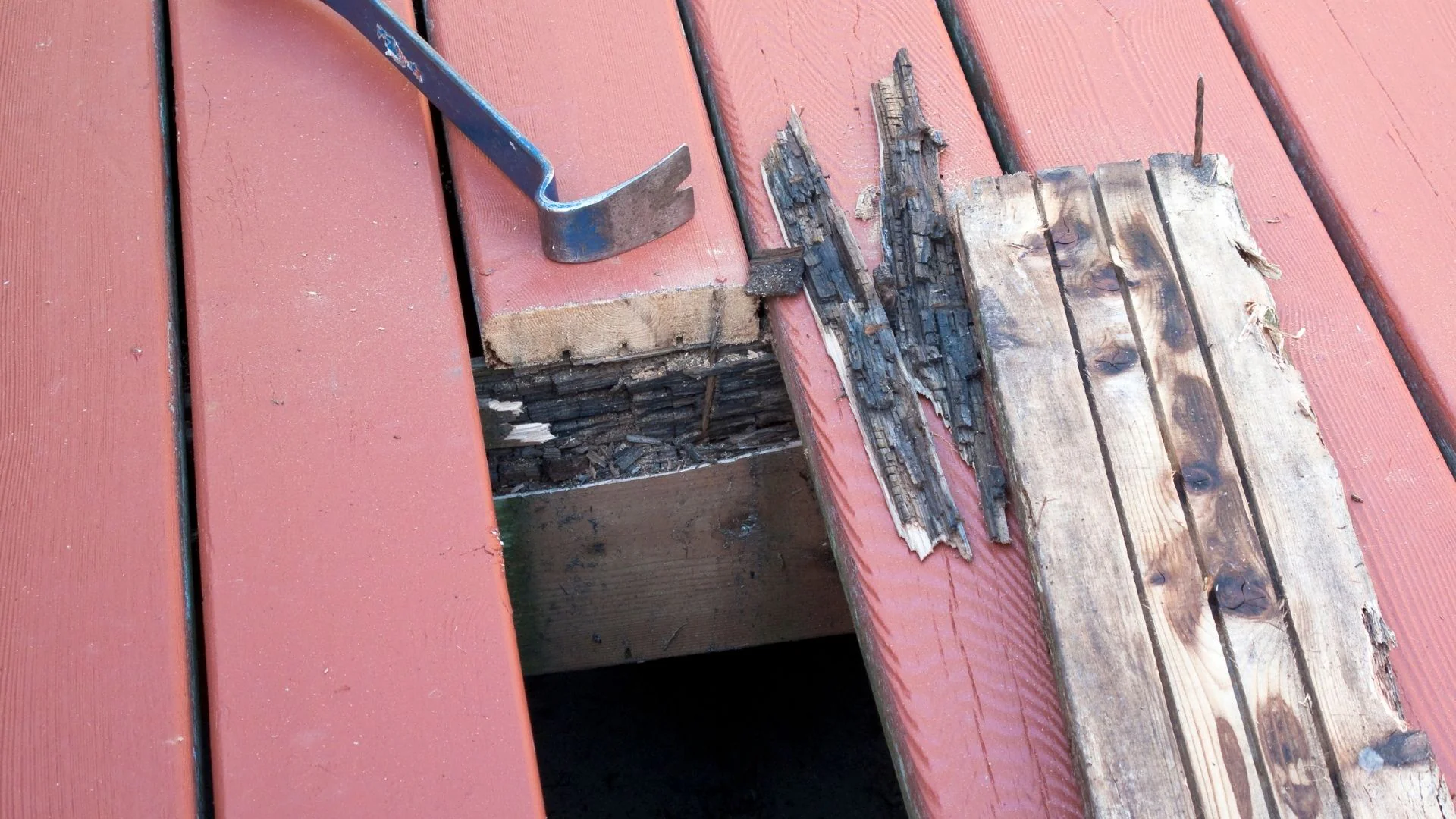 A pry bar is placed next to rotting deck wood being removed from a red-painted surface during repair.
