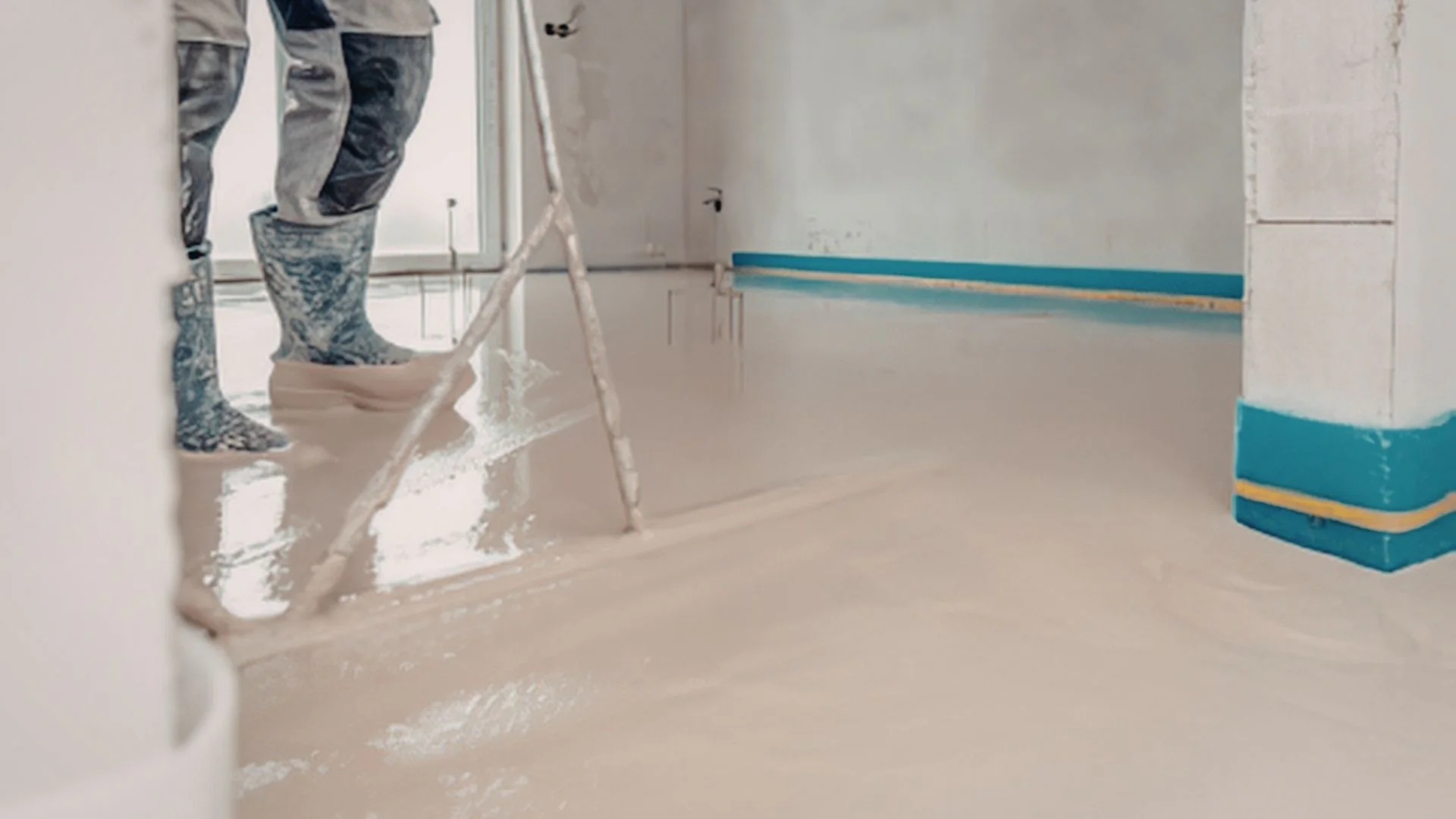 A person in boots uses a tool to spread self-leveling liquid floor across a room’s surface.