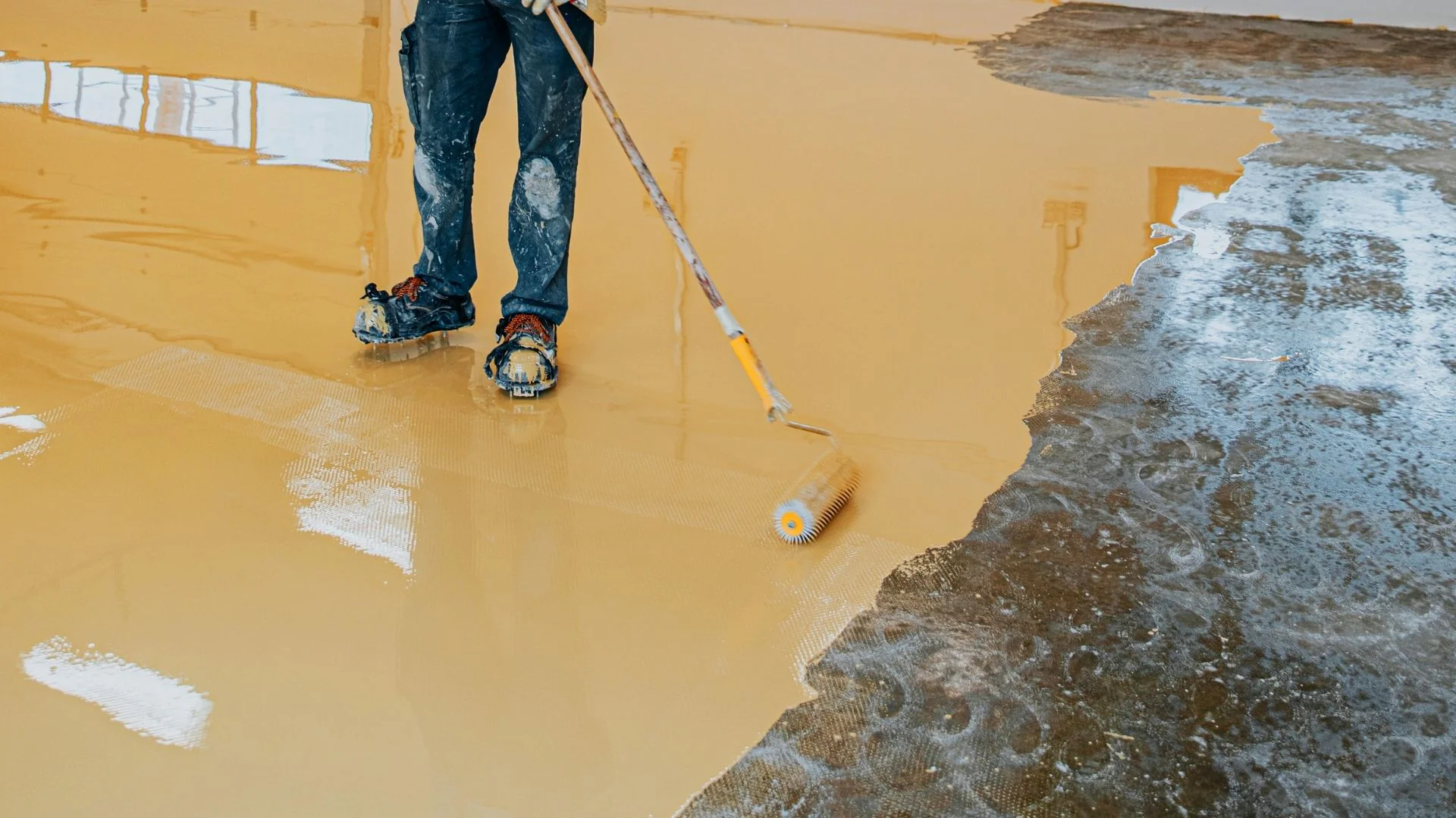 A person applies beige epoxy coating with a paint roller to a concrete floor in an indoor area.