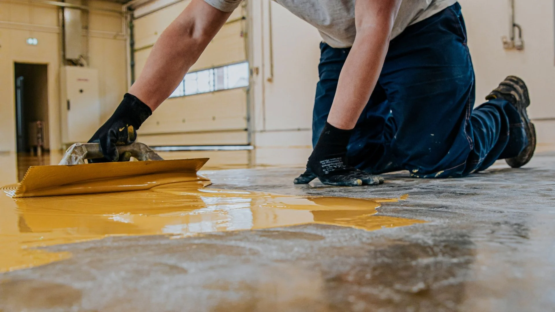A person in gloves uses a trowel to spread yellow epoxy paint on a concrete garage workshop floor.
