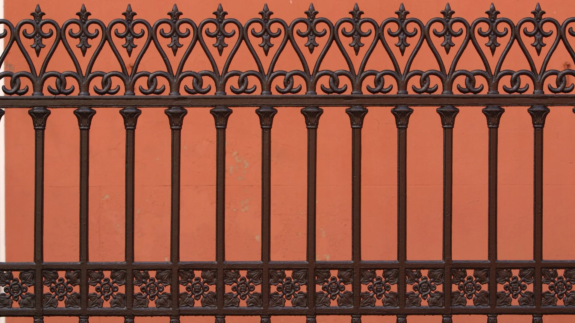 An ornate black wrought iron fence with fleur-de-lis designs stands before a vibrant orange wall background.