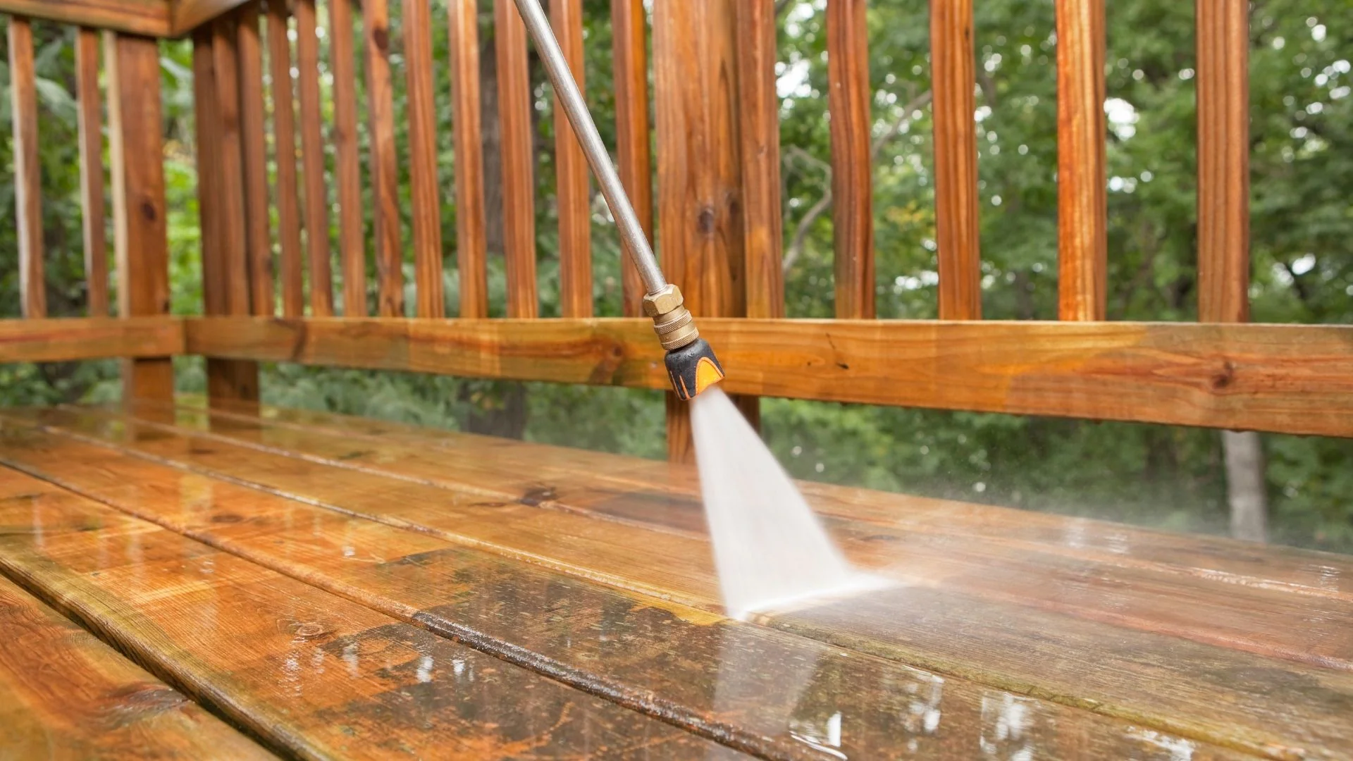 A pressure washer cleans dirt from a wooden deck, with water spraying near a wood railing in the background.