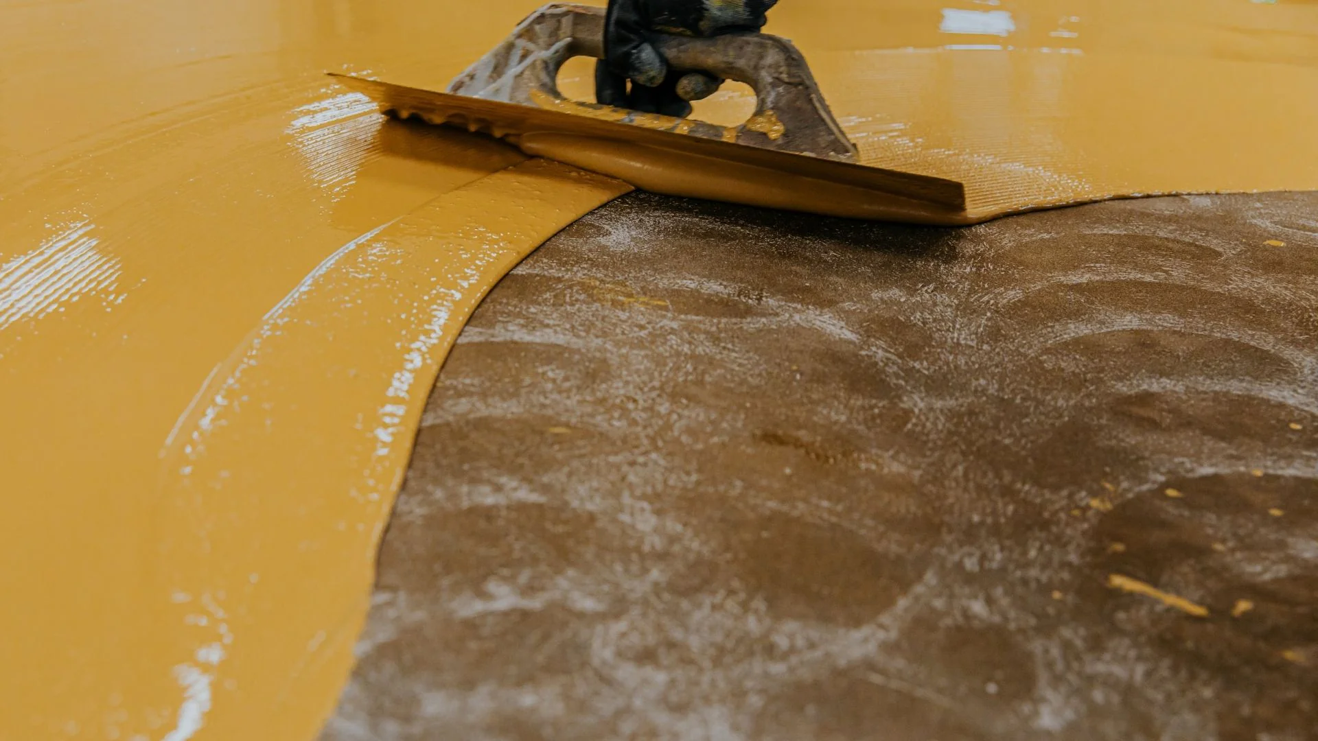 A gloved hand uses a notched trowel to spread yellow floor adhesive onto a textured flooring surface.